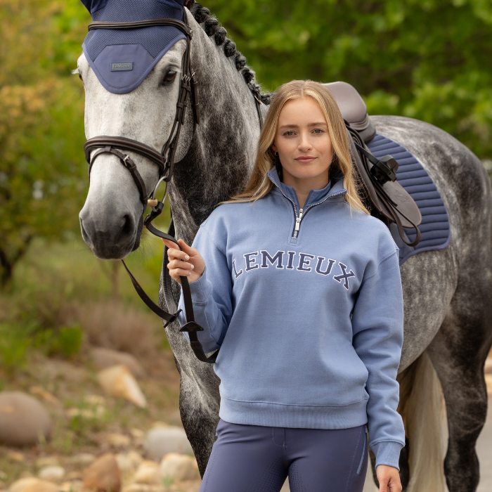 Woman wears a blue sweater with LEMIEUX logo in an outdoor setting with a horse
