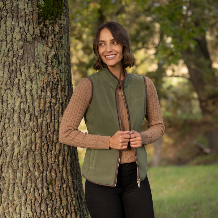 LeMieux AW26 Frankie Fleece Gilet Variant Colour Willow Green.
A woman stands outdoors next to a large tree trunk, wearing an willow green zip-front vest over a beige cable-knit sweater and black pants.