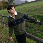 LeMieux AW26 Mens Rugby Shirt Varient Olive. A man wearing a Lemieux long-sleeve rugby shirt in olive green and navy blue with a white collar, standing outdoors in a grassy field next to a wooden fence.