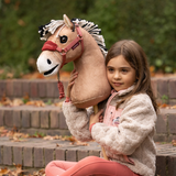LeMieux AW26 Hobby Horse Oskar Variant Colour Brown.
A young girl with brown hair sits on outdoor brick steps holding a plush toy horse. The toy horse has a tan body, a white and black mane, and a pink halter.