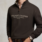 Holland Cooper Heritage Quarter Zip Sweat (Men's) Variant Bournvile. Man wearing a brown 1/4 zip sweatshirt with Holland Cooper branding across the chest in white agaisnt a grey background.