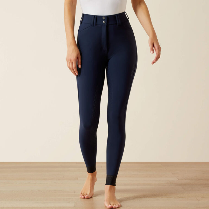A model wearing navy blue equestrian riding breeches, standing barefoot on a light wood floor against a warm beige background.