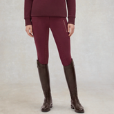 A model wearing tight equestrian riding breeches in deep burgundy, paired with a matching long-sleeve top and dark brown riding boots, standing against a neutral studio backdrop.