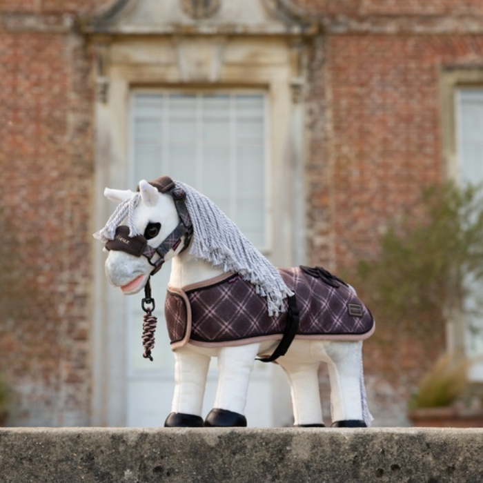 LeMieux AW26 Toy Pony Heritage Rug Varient Cocoa. A small white plush toy horse wearing a dark plaid saddle blanket and a gray fringed mane, posed outdoors in front of a blurred brick building.