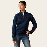 Ariat Youth Team 1/2 Zip Pullover Variant Navy. A woman modeling a navy blue quarter-zip pullover sweatshirt with a small red embroidered logo on the left chest and red text on the right sleeve, paired with blue jeans against a light beige background.