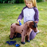 LeMieux Toy Pony Variant Gracie. A young girl kneeling on a green grass lawn, playing with a brown plush toy horse. She is wearing a purple and white raglan shirt with a unicorn graphic and matching purple leggings.