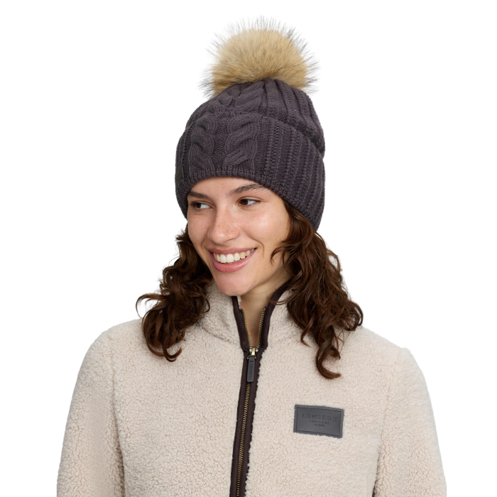 A woman wearing a dark gray cable-knit beanie with a beige faux-fur pom-pom, paired with a cream fleece jacket, against a solid white background.
