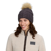A woman wearing a dark gray cable-knit beanie with a beige faux-fur pom-pom, paired with a cream fleece jacket, against a solid white background.