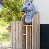 LeMieux AW26 Hobby Horse Stable Wooden Variant Colour Brown.
A gray horse-shaped plush or stuffed toy sits inside a tall wooden cabinet with a slatted front, photographed outdoors against a white brick wall and greenery.