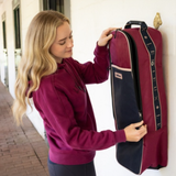 LeMieux AW26 Anatomical Bridle Bag Varient Blackberry. A woman in a maroon hoodie is shown from the side, holding a tall, zippered storage bag or organizer against a white wall. The bag is burgundy and navy blue with a vertical black stripe featuring gold lettering that reads 'LeMieux'.