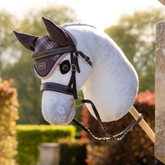 LeMieux AW26 Hobby Horse Heritage Fly Hood Varient Cocoa. A white hobby horse wearing a dark brown plaid fly veil and black bridle, mounted on a wooden pole against a blurred garden background.