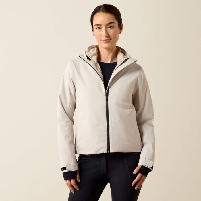 A woman modeling a light beige hooded zip-up jacket with black trim, worn over a dark navy top and black pants, against a plain beige studio background.
