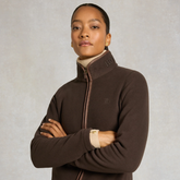 Holland Cooper Country Fleece Jacket Variant Chocolate. A woman wearing a dark brown zip-up fleece jacket with a high collar and a cream ribbed turtleneck underneath, standing with arms crossed against a textured light gray background.
