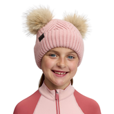 LeMieux AW26 Mini Double Pom Beanie Variant Petal. A smiling young girl wearing a light pink ribbed knit beanie with two large beige faux-fur pom-poms and a small black logo tab, paired with a pink and coral zip-up top against a plain white background.