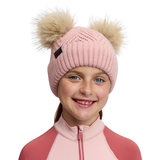 LeMieux AW26 Mini Double Pom Beanie Variant Petal. A smiling young girl wearing a light pink ribbed knit beanie with two large beige faux-fur pom-poms and a small black logo tab, paired with a pink and coral zip-up top against a plain white background.