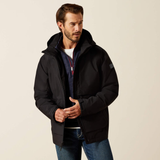 A man wearing a black hooded puffer jacket over a navy and white quarter-zip sweater and blue jeans, standing against a plain light beige studio background.