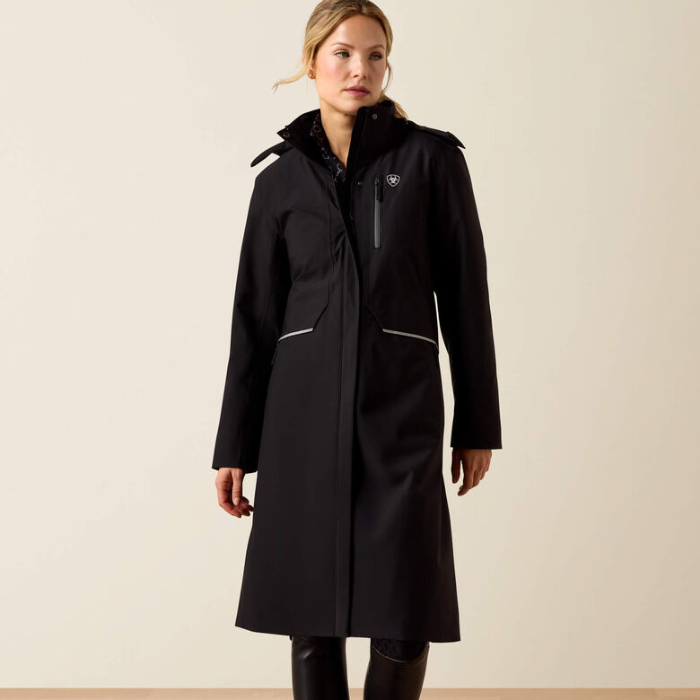 A woman modeling a long black hooded coat with reflective silver trim and a small shield logo on the chest, photographed against a plain beige studio background.