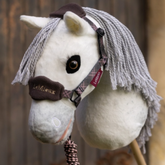 LeMieux AW26 Hobby Horse Heritage Vogue Headcollar & Leadrope Varient Cocoa. A plush toy horse head with a white face, gray yarn mane, and dark brown velvet eye cover, wearing a plaid LeMieux halter against a blurred wooden background.