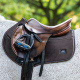 Woof Wear Elite Comfort Jump Saddle Pad Variant Chocolate. A brown leather saddle with black stirrups and straps, resting on a dark brown saddle pad on the back of a horse, with a blurred green outdoor background.