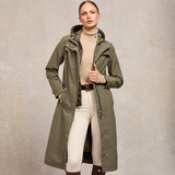 Holland Cooper Folgate Rain Coat Variant Soft Khaki. Woman wearing a long khaki coat with gold button detailing over a beige jumper and white jeans against a grey background.