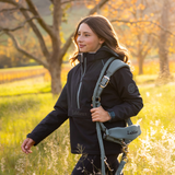 LeMieux AW26 Young Rider Bailey Waterproof Anorak Varient Navy. A woman walking outdoors in a grassy field at golden hour, wearing a navy LeMieux jacket and carrying a gray LeMieux horse halter and lead strap over her shoulder.