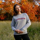 LeMieux AW26 Vida Varsity Crew Neck Sweat Variant Colour Grey Marl.
A woman wearing a light gray crewneck sweatshirt with 'LEMIEUX' printed in red across the chest, standing in a grassy field with autumn trees in the background.