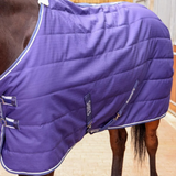 A close-up of a horse wearing a purple quilted horse blanket with white piping and silver buckles. The brand name 'SHIRES' is visible on a dark blue label sewn onto the side of the blanket.