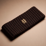 A dark brown ribbed knit headband with a small gold interlocking monogram logo in the center, displayed on a neutral beige background.