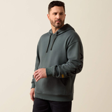 A man wearing a dark slate green pullover hoodie with black drawstrings and black graphic lettering on the sleeve, standing against a plain beige background.