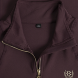 A Holland Cooper Cranborne Long Sleeve Base Layer Variant Colour Mulberry.
Close-up of a dark plum purple quarter-zip pullover jacket, showing a gold-tone zipper, a black XS size tag, and a gold embroidered crest logo on the lower right.