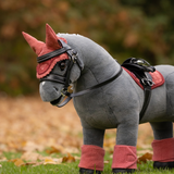 LeMieux AW26 Toy Pony Fly Hood Variant Colour Spiced Earth.
A plush toy horse wearing a spiced earth colour equestrian costume, including a horned bridle, black faux-fur mane, saddle, and leg wraps, standing on grass with a blurred outdoor background