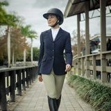 Person in equestrian attire walking on a path with trees and a building in the background