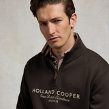 Holland Cooper Heritage Quarter Zip Sweat (Men's) Variant Bournville. Close up of man wearing a 1/4 zip with white Holland Cooper branding and brown zip detaling against a grey background.