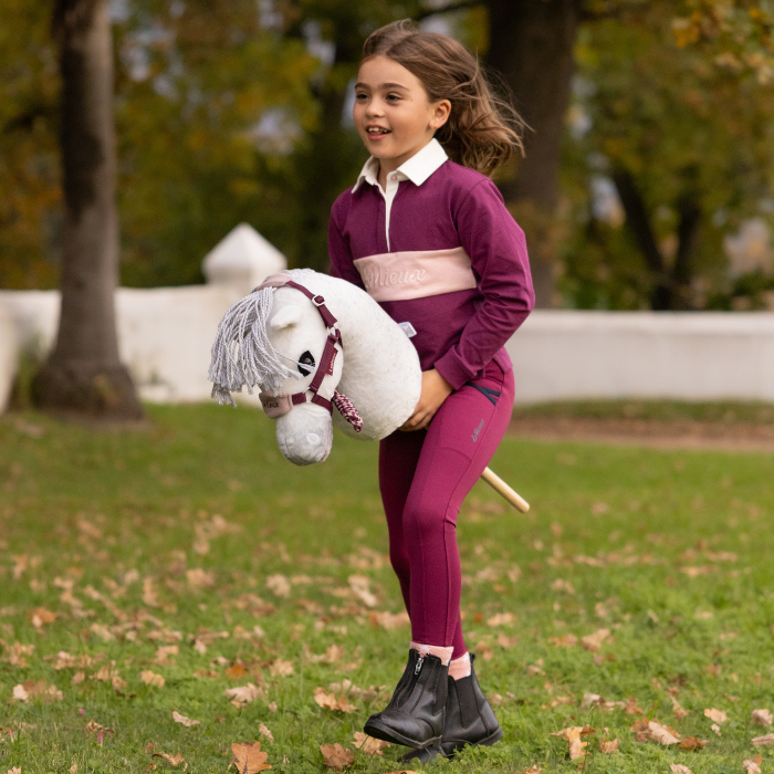 LeMieux AW26 Hobby Horse Coco Variant Colour White.
A young girl stands on a grassy park lawn wearing a matching burgundy and pink equestrian outfit, holding a plush white hobby horse toy.