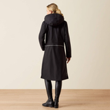 A woman viewed from the back wearing a long black hooded coat, black leather boots, and black leggings, standing on a light wood floor against a plain beige wall.