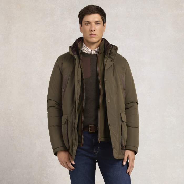 Holland Cooper Stamford Field Coat (Men's) Variant khaki. A man wearing an olive green hooded puffer jacket over a dark sweater and blue jeans, standing against a textured light gray studio background.