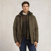 Holland Cooper Stamford Field Coat (Men's) Variant khaki. A man wearing an olive green hooded puffer jacket over a dark sweater and blue jeans, standing against a textured light gray studio background.