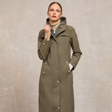 Holland Cooper Folgate Rain Coat Variant Soft Khaki. Woman wearing a long khaki zipped up coat with gold button detailing against a grey background.