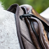 Woof Wear Elite Comfort Jump Saddle Pad Variant Chocolate. Close-up of a brown leather riding saddle with a black strap and textured brown pad, resting on a white horse with a blurred green outdoor background.