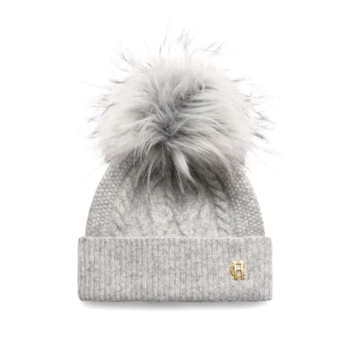 Holland Cooper Sloane Cable Bobble Hat varient Grey Marl. A light gray cable-knit beanie with a large fluffy gray pom-pom and a small gold metal logo charm on the folded cuff, shown against a plain white background.