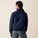 A woman shown from the back wearing a navy blue bomber jacket and blue jeans against a plain light beige studio background.