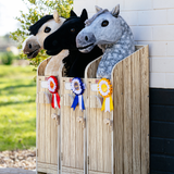 LeMieux AW26 Hobby Horse Stable Wooden Variant Colour Brown.
Three plush horse heads in a wooden display stand outdoors, with rosettes attached in front. The horses are cream, black, and dappled gray, set against a background of a white brick wall and green grass.
