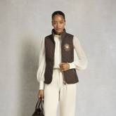 Holland Cooper Boughton Diamond Quilt Gilet Variant Colour Walnut.
A model stands against a textured gray studio backdrop wearing a dark brown quilted zip-front vest over a cream long-sleeve top and matching cream trousers. The vest has a small embroidered crest on the left chest.
