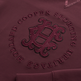 Holland Cooper Cranborne Crystal Crew Variant Mulberry. Close-up of a maroon hoodie featuring an embossed circular design with the text 'HOLLAND COOPER SPORTING HERITAGE' and a large decorative monogram in the center.