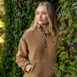 LeMieux AW26 Avery Teddy Quarter Zip Fleece Varient Caramel. A woman with blonde hair wearing a brown sherpa fleece quarter-zip pullover jacket, standing outdoors in front of a green leafy hedge.