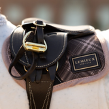 LeMieux AW26 Toy Pony Heritage Pad Varient Cocoa. A close-up of a black leather equestrian saddle with gold-tone hardware and a plaid-patterned saddle pad. A black leather brand patch reading 'LEMIEUX HERITAGE' is visible on the side.