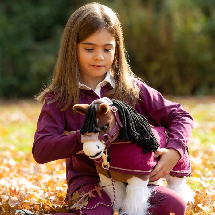 LeMieux AW26 Toy Pony Variant Colour Titan. A young person sat on grass holding a toy plush pony, all wearing blackberry colour items. on faded garden background.