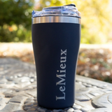 LeMieux AW26 Travel Cup Varient Navy. A dark navy blue insulated travel tumbler with a clear lid and a silver stainless steel rim, featuring the name 'LeMieux' printed vertically in white. The tumbler is photographed outdoors on a stone surface with a blurred natural background.