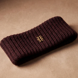 A dark burgundy ribbed knit headband with a small gold interlocking monogram logo, displayed on a textured beige surface.