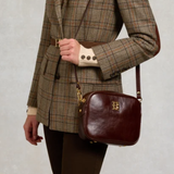 Holland Cooper Country Camera Bag Variant Chesnut Leather. Person wearing a tweed jacket with a leather chesnut crossbody bag with gold detailing.
